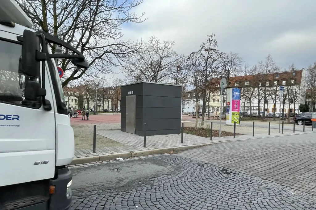 Eine graue Toilettenanlage steht neben einer Kopfsteinpflasterstraße, die von kahlen Bäumen in der ruhigen Stadt gesäumt ist. Links ist ein weißer Lastwagen teilweise sichtbar, während im Hintergrund Reihen von Häusern und bunte Schilder unter einem bewölkten Himmel hervorschauen.