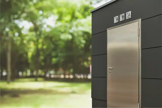 Eine moderne öffentliche Toilette mit einer Metalltür steht vor einem verschwommenen Hintergrund aus üppigen, grünen Bäumen. Symbole über der Tür weisen auf Einrichtungen für Männer, Frauen und Menschen mit Behinderungen hin.