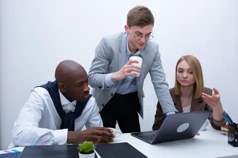 Auf dem Foto ist eine diverse Gruppe von 3 Menschen abgebildet. Sie sitzen und stehen vor einem Laptop und einem Stapel Papier an einem Tisch.