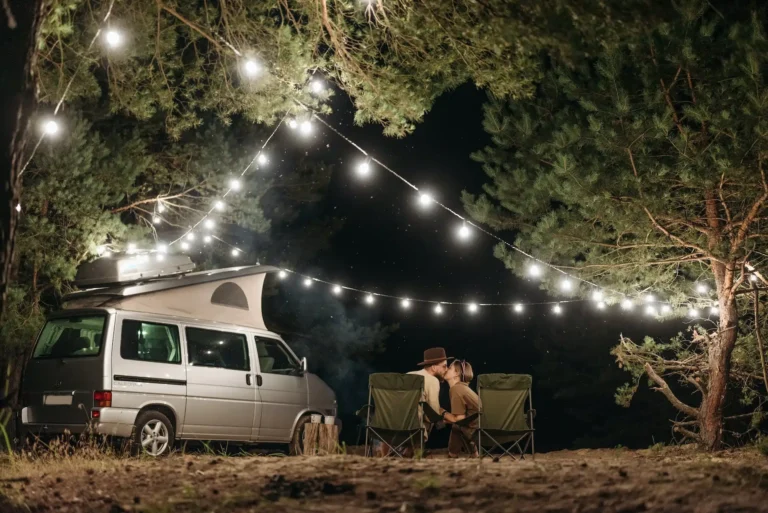 Ein Camper in der Nacht neben einem küssenden Pärchen, umgeben von Bäumen, an denen Lichterketten aufgespannt sind