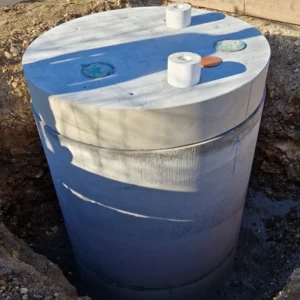 Die TTB GOLDGRUBE Betongrube ist auf einer Baustelle teilweise eingegraben und von Erde und Holzbalken umgeben. Oben ist der kreisförmige Deckel mit fünf Löchern und Deckeln zu sehen.