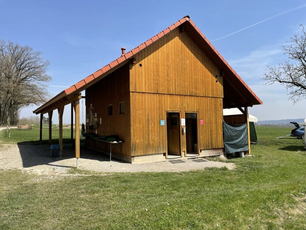 Ein Holzgebäude mit rotem Dach beherbergt die Toiletten für Männer und Frauen. Es ist von Gras umgeben, mit einem Baum und einer offenen Landschaft im Hintergrund.