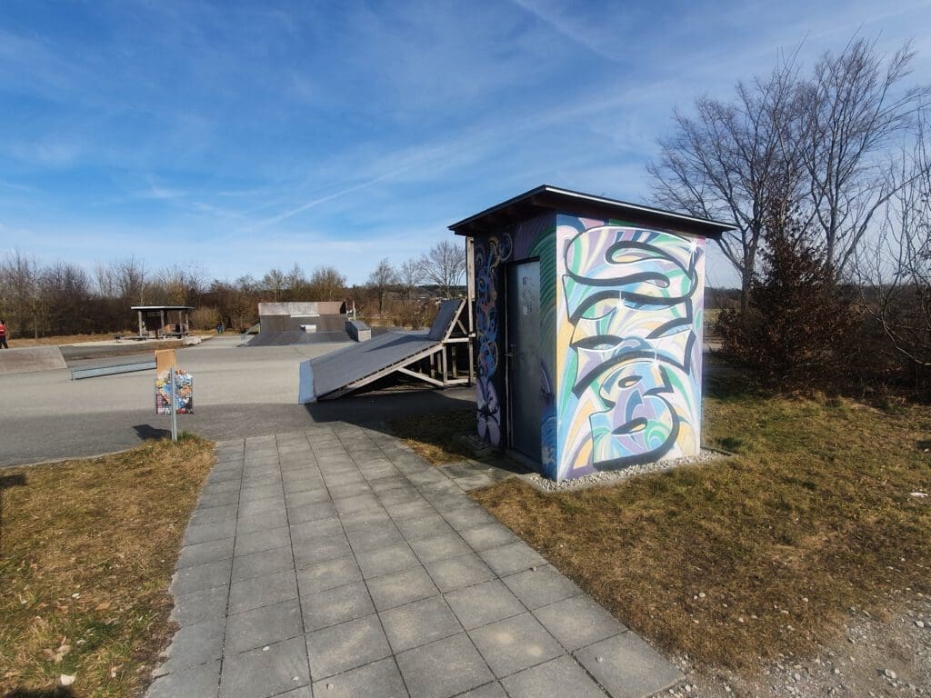 Ein Skatepark unter einem klaren blauen Himmel bietet Rampen und mit Graffiti bedeckte Strukturen. Ein gepflasterter Weg führt zu einer farbenfrohe Toilettenanlage mit abstrakter Kunst. Bäume und offene Felder umgeben das Gebiet.