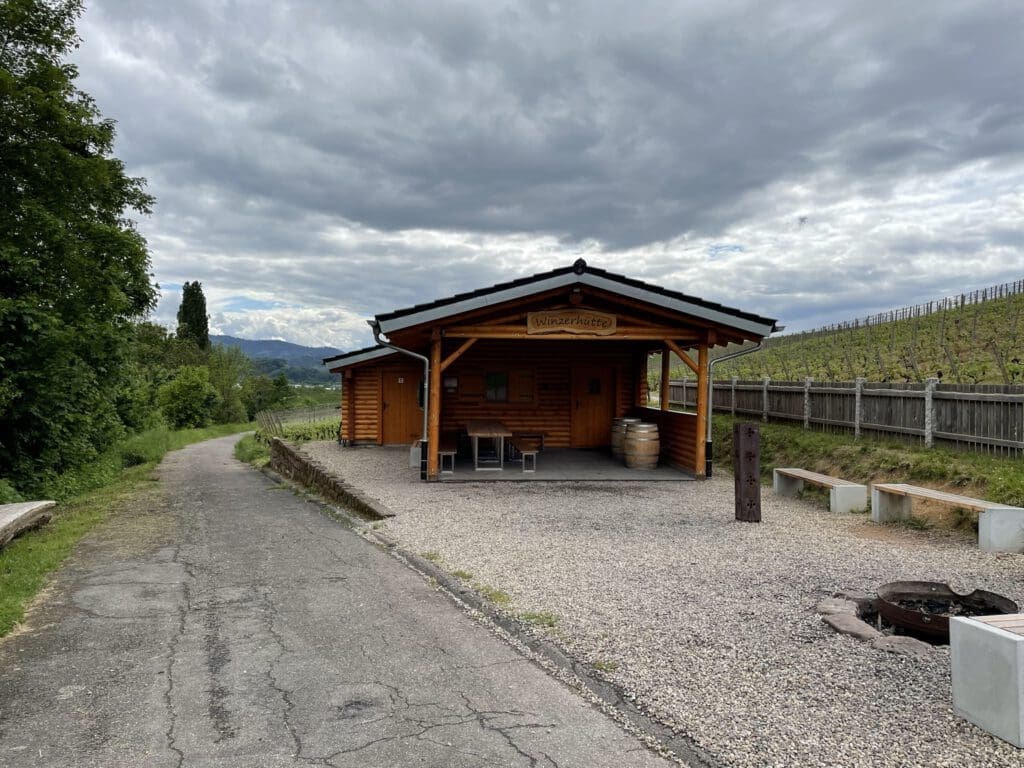 Eine kleine Holzhütte, die einer traditionellen Grillhütte mit überdachter Veranda ähnelt, steht an einem Kiesweg. Im Vordergrund stehen zwei Bänke und eine leere Feuerstelle. Rechts erstreckt sich unter dem wolkenverhangenen Himmel ein Weinberg entlang eines Hügels.