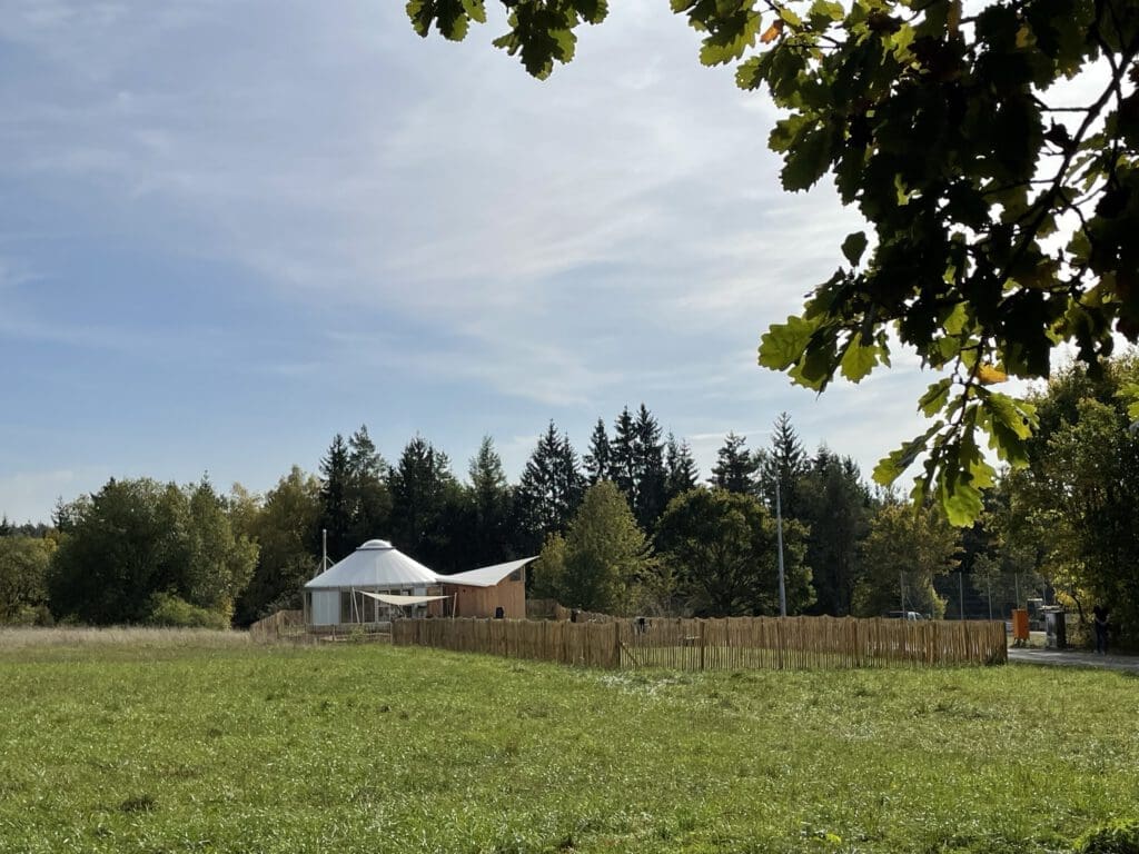 Eine Jurte mit einem Holzzaun steht auf einer Wiese unter blauem Himmel und erinnert an einen Waldkindergarten. Bäume säumen das Gelände, und große Blätter rahmen die Szene von oben ein und schaffen eine friedliche und natürliche Landschaft.