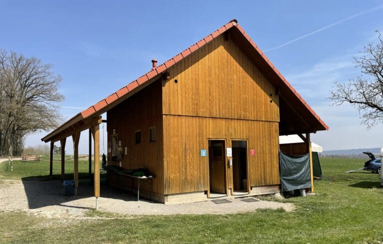 Ein Holzgebäude mit rotem Dach beherbergt die Toiletten für Männer und Frauen. Es ist von Gras umgeben, mit einem Baum und einer offenen Landschaft im Hintergrund.