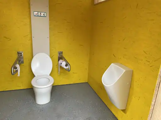 Ein Toilettenraum mit gelben Wänden verfügt über eine Toilette, ein Urinal und zwei an der Wand montierte Halter für Toilettenpapier und Stützen für eine barrierefreie Umgebung. Die Trockentrenntoilette sorgt für eine barrierefreie und nachhaltige Nutzung.