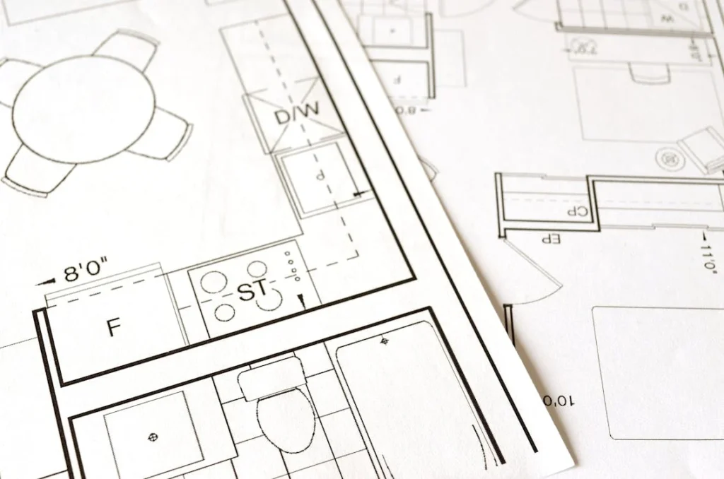 Eine Nahaufnahme architektonischer Pläne, die einem komplizierten Datenblatt ähneln, zeigt einen Grundriss einer Küche mit Herd und Spüle sowie eines Badezimmers mit Toilette. Die Pläne sind elegant über eine makellos weiße Oberfläche verteilt.