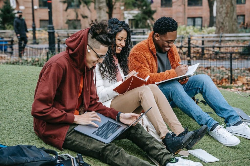 Studenten sitzen auf einem Grashügel und haben verschiedene Medien in der Hand, wo sie Informationen nachlesen. Das wollen wir auch tun. Sie sind auf der Suche nach mehr Wissen.