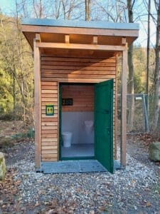 Auf dem Foto ist eine Toilette auf einem Parkplatz im Wald zu erkennen. Ihre grüne Türe ist offen und man kann Urinal und Toilettenstuhl erkennen.