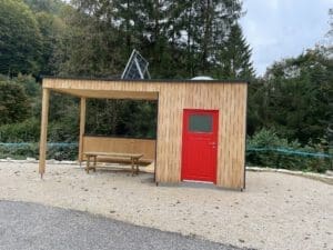 Eine kleine autarke Toilettenanlage aus Holz mit offenem Bereich und leuchtend roter Tür im Freien. Sie verfügt über einen Picknicktisch unter dem Unterstand und ein Solarpanel auf dem Dach, das Energieunabhängigkeit signalisiert. Umgeben von grünen Bäumen und einem bewölkten Himmel im Hintergrund.