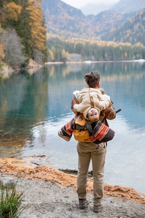 In der Mitte des Bildes ist ein Vater mit einem Kind auf dem Rücken. Das Kind hängt kopfüber auf den Schultern und dem Rucksack und lacht. Im Hintergrund ist ein See und Berge voll grün-gelber Bäume im Herbst.