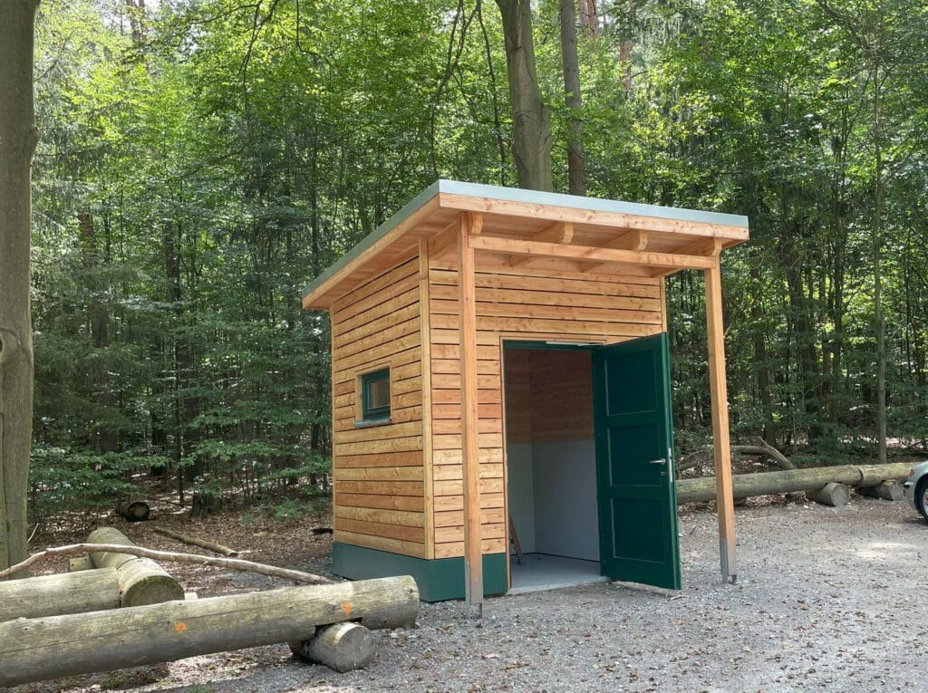 In der Mitte des Fotos ist eine Holzhütte, ein Toilettenhaus. Im Vordergrund ist eine Holzbank zum Hinsetzen und im Hintergrund viel grün von verschiedenen Bäumen. Das Bild dient auch als Referenz für unsere Sanitäranlage im Osten.