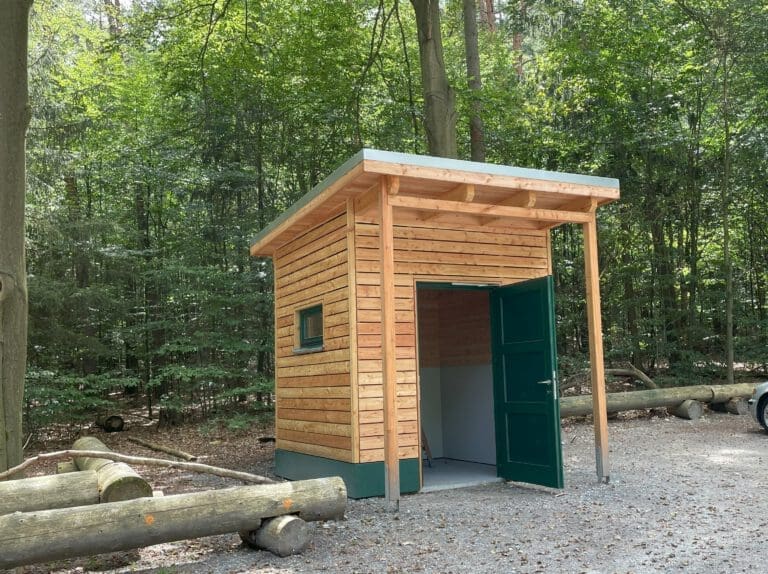 In der Mitte des Fotos ist eine Holzhütte, ein Toilettenhaus. Im Vordergrund ist eine Holzbank zum Hinsetzen und im Hintergrund viel grün von verschiedenen Bäumen