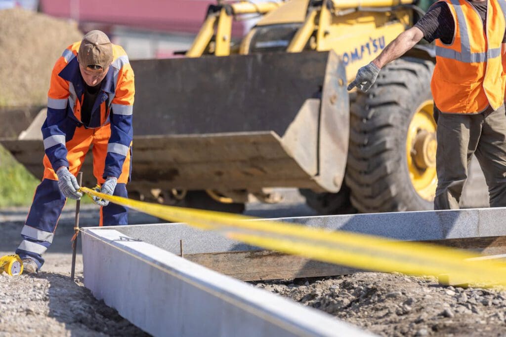 Im Vordergrund ist eine Grube. Ein Bauarbeiter hängt gerade ein Flatterband um die Grube auf. Im Hintergrund sieht man einen Radlader und einen weiteren Bauarbeiter.