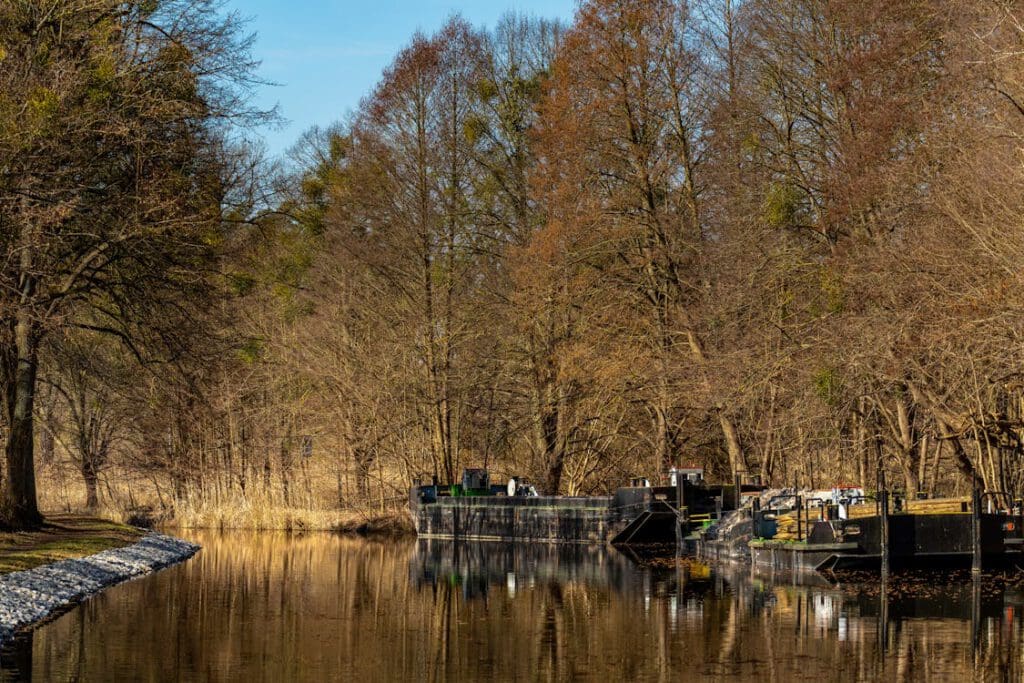 Durch die Mitte des Bildes fließt ein Fluss. An seinem rechten Ufer liegen zwei alte, kleine Kähne an. Im Hintergrund dieser Kähne sowie am linken Ufer sieht man überall Bäume im Herbst mit wenigen Blättern.