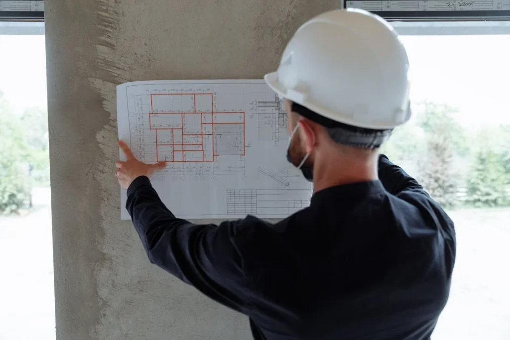 Ein Bauarbeiter mit einem weißen Helm hält einen Plan mit orangefarbenen Quadraten gegen eine Wand.