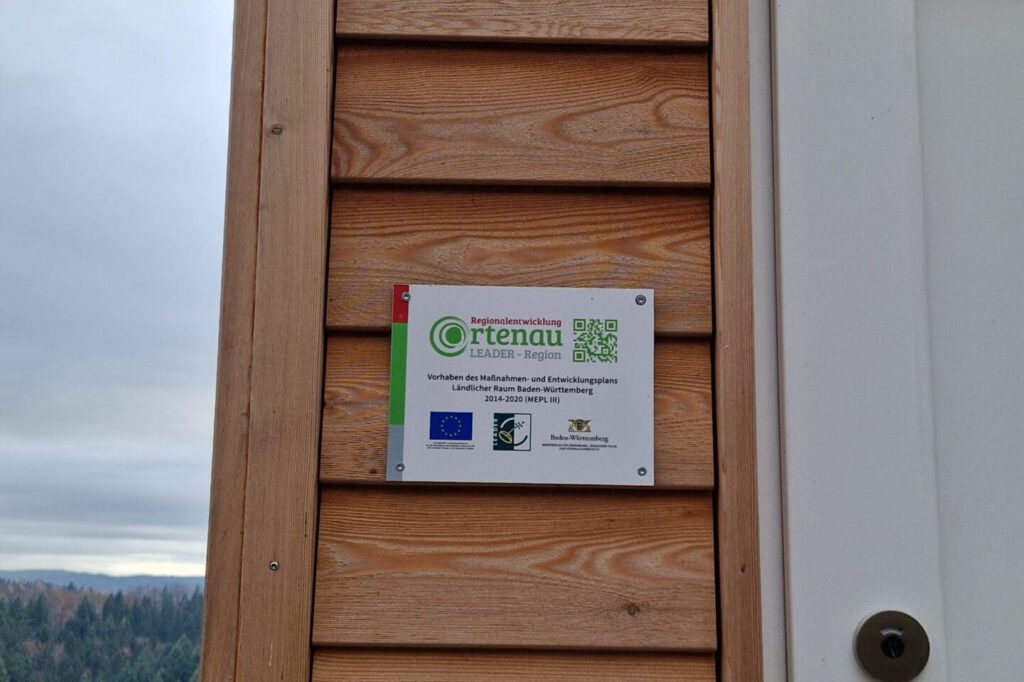 Ein Schild hängt an einer Holzwand neben einer Türe. Auf dem Schild steht Regionalentwicklung Ortenau LEADER-Region und beschreibt ein Förderprogramm der EU. Eine Förderung ist auch für Ihre Toilette möglich.