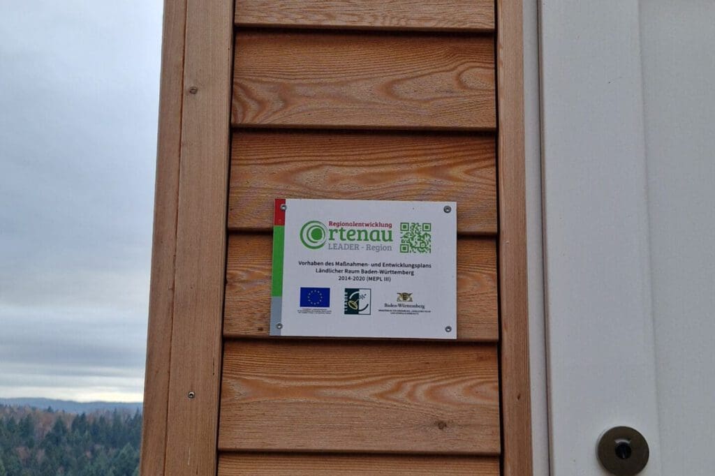 Ein Schild hängt an einer Holzwand neben einer Türe. Auf dem Schild steht Regionalentwicklung Ortenau LEADER-Region und beschreibt ein Förderprogramm der EU.