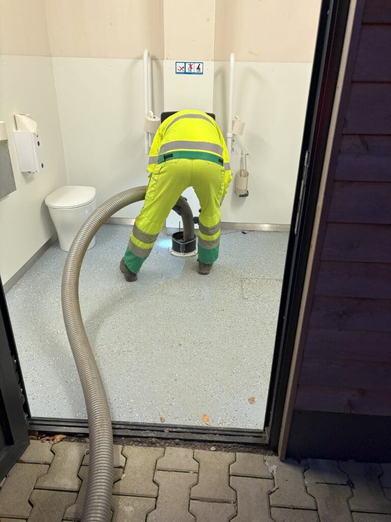 Ein Mann mit einem neongelben Schutzanzug hat einen Absaugschlauch in einem Toilettenraum in der Hand. Er entleert gerade eine GOLDGRUBE. Der Toilettenstuhl steht abgebaut links im Eck.