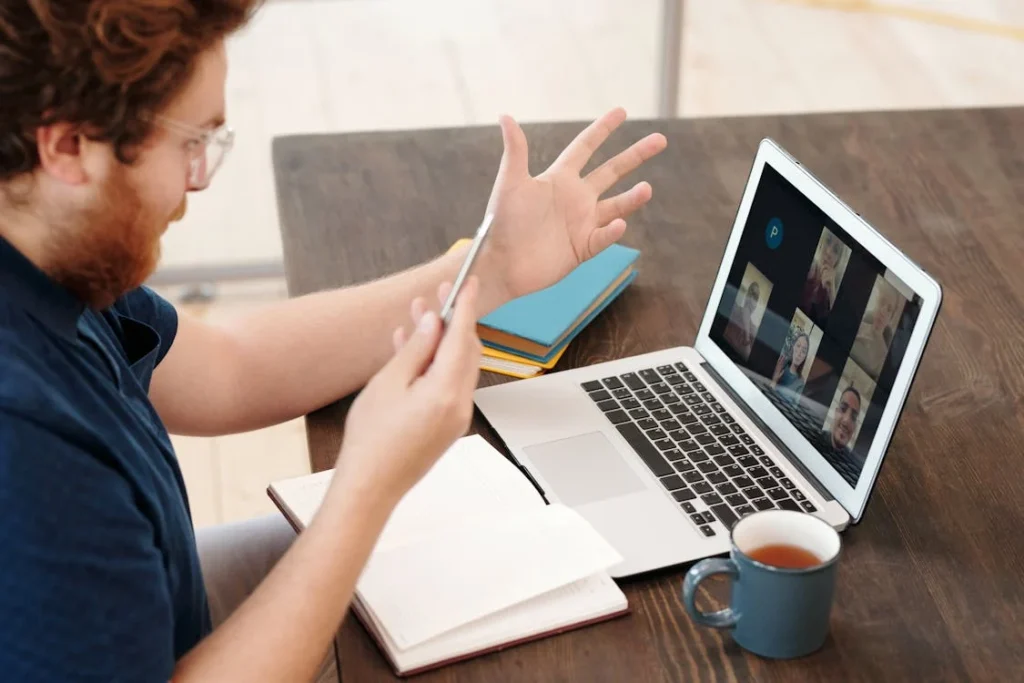 Auf dem Foto ist ein Mann, der vor einem Laptop an einem Holztisch sitzt. Auf dem Laptop sind verschiedene Leuten zu sehen, mit denen er sich unterhält. Vor Ihm liegt außerdem ein offenes Buch und es steht eine blaue Tasse mit Tee daneben.