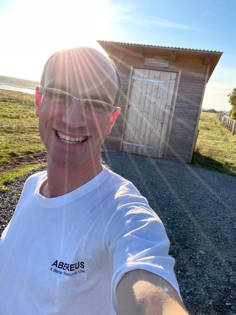 Auf dem Foto ist unser Erfinder Flo mit einem ABEREUS T-Shirt an und einer unserer Anlagen im Hintergrund. Es scheint die Sonne mit blauem Himmel.