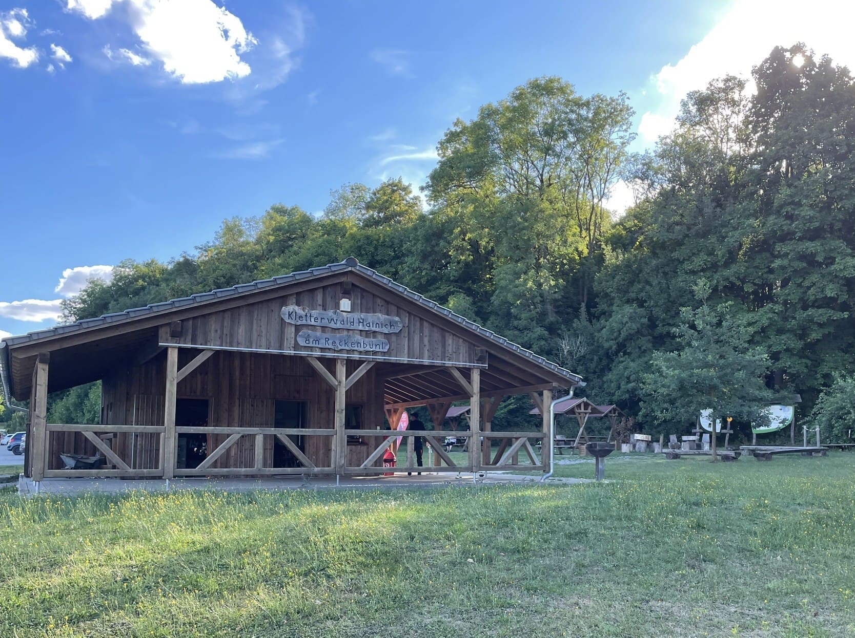 Auf dem Foto ist ein Holzhaus im Vordergrund und im Hintergrund sieht man einen Wald und Sitzmöglichkeiten aus Holz. Auf dem Haus steht Kletterwald Hainich. Der Himmel ist blau und die Wiese im Vordergrund saftig grün.