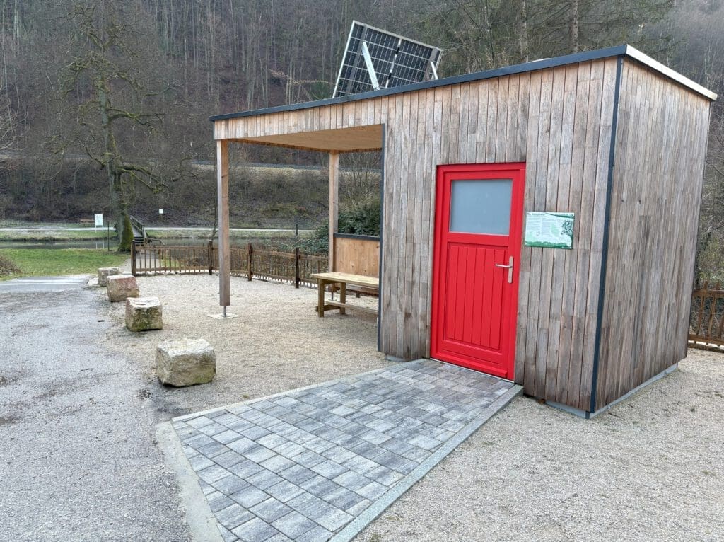 Eine kleine autarke Toilettenanlage aus Holz mit offenem Bereich und leuchtend roter Tür im Freien. Sie verfügt über einen Picknicktisch unter dem Unterstand und ein Solarpanel auf dem Dach, das Energieunabhängigkeit signalisiert. Umgeben von grünen Bäumen und einem bewölkten Himmel im Hintergrund.