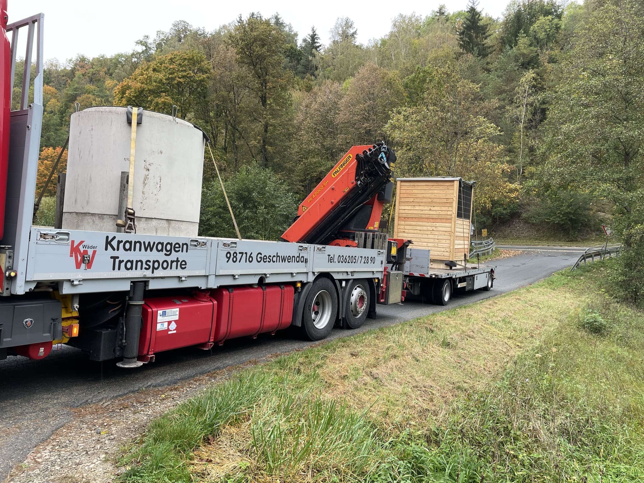 Auf dem Foto ist ein LKW mit einer Goldgrube drauf. Dieser hat auch einen Anhänger mit einem Toilettenhaus aus Holz drauf. Der Laster steht auf einer kleinen Straße mit Wiesen und Wäldern im außen herum