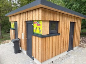 Auf dem Foto ist eine Toilettenanlage. Auf der linken Seite sind die Eingänge zu der Herren und Damentoilette mit jeweils grauen Türen. Dazwischen steht eine graue Mülltonne. Rechts ist der Eingang zu einem Extraraum. Das Haus ist aus Holz und die Toilettenschilder sind bunt und es sind Bienen abgebildet.