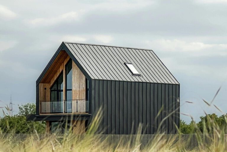Auf dem Foto ist ein Tiny Haus in der Natur. Im Vordergrund ist Getreide auf eine Feld und in der Mitte des Bildes ein Haus. Es handelt sich um ein graues Tiny House mit einer Fensterfront auf der einen kleinen Seite und eine fensterlose, größere Wand. Der Himmel ist grau und mit Wolken bedeckt.