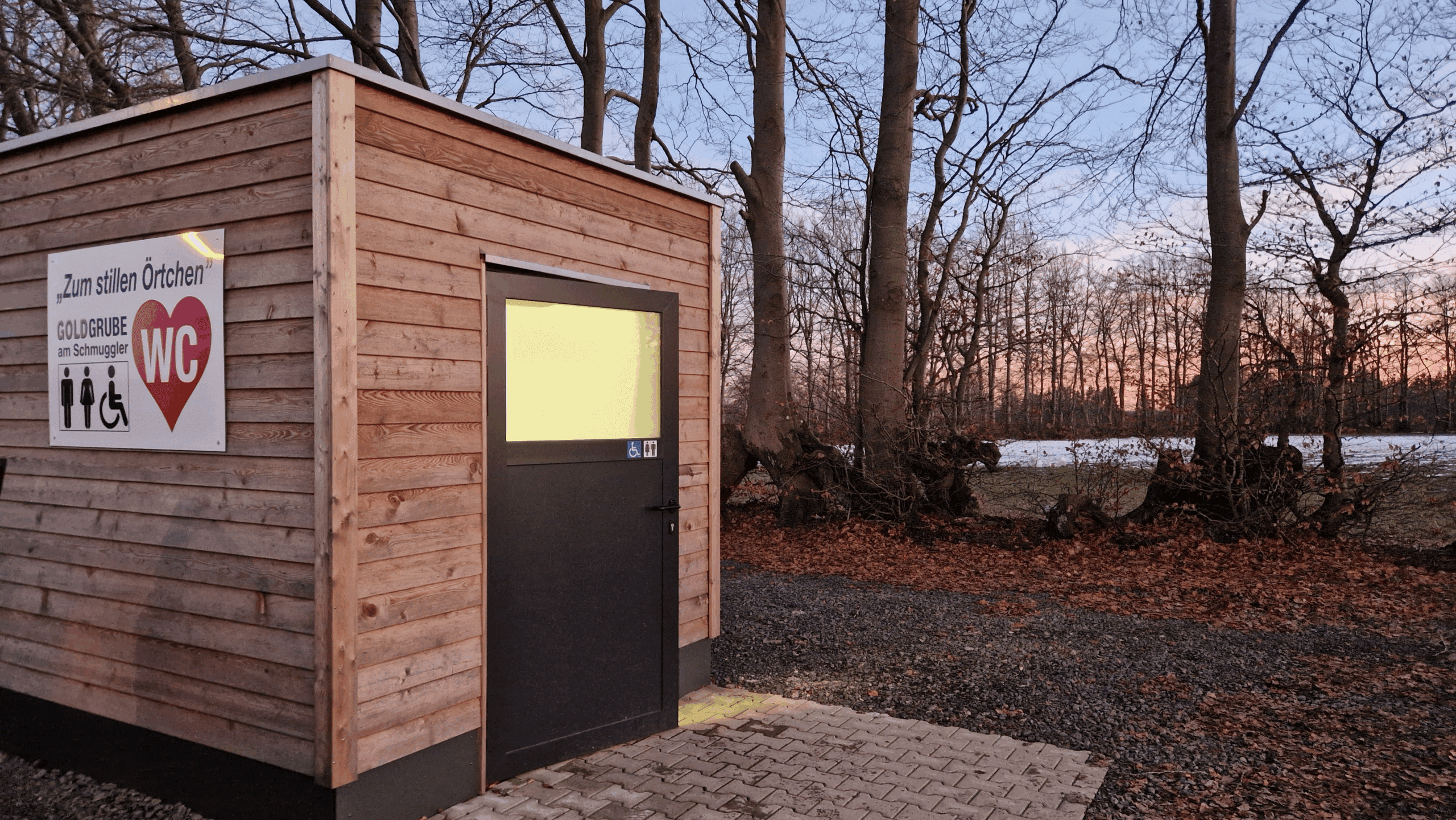 Das Foto zeigt ein Toilettenhäuschen in dem Licht durch das Milchglasfenster der Türe scheint. Das Haus steht am Rande eines Parkplatzes mit Bäumen und einer Wiese im Winter im Hintergrund. Die Sonne geht gerade unter und in der Ferne sieht man das Abendrot.