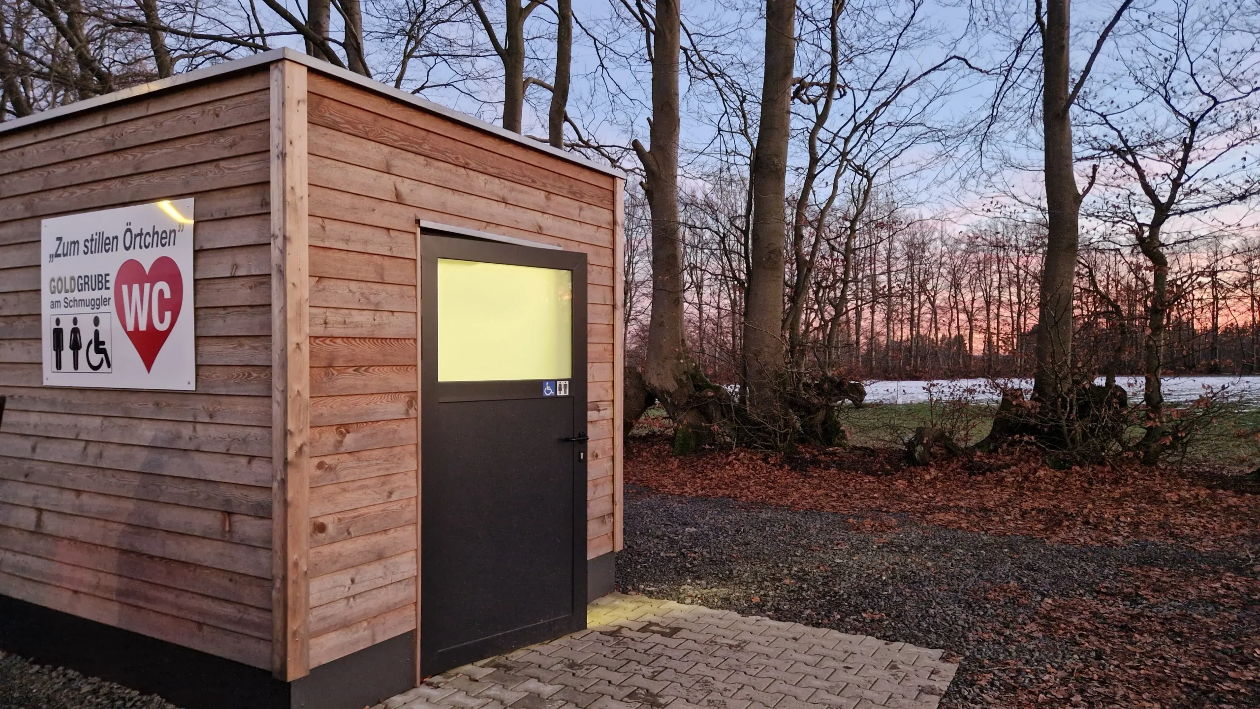 Das Foto zeigt ein Toilettenhäuschen in dem Licht durch das Milchglasfenster der Türe scheint. Das Haus steht am Rande eines Parkplatzes mit Bäumen und einer Wiese im Winter im Hintergrund. Die Sonne geht gerade unter und in der Ferne sieht man das Abendrot.
