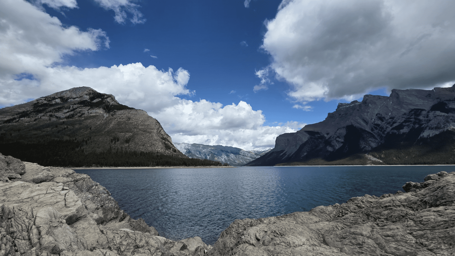 Auf dem Foto ist ein See in den Bergen. Wolken spiegeln sich auf dem blauen Wasser.