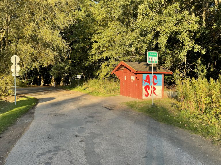 Auf dem Foto ist eine alte Toilettenanlage in Rostock zu erkennen. Das rote Toilettenhaus steht an einer kleinen Straße und ist seitlich beschmiert. Dahinter ist ein dichter Wald zu erkennen.