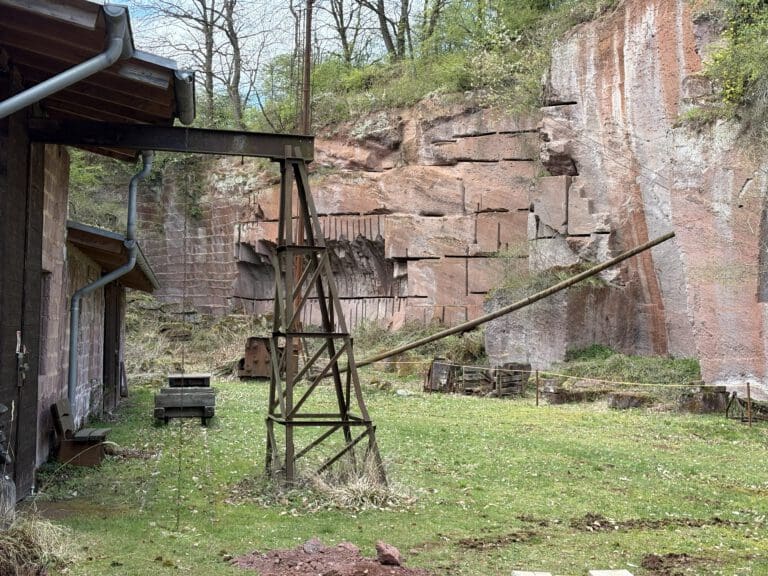 Auf dem Foto ist der Steinbruch Michelnau. Im Hintergrund sind rote Felswände und davor verschiedene Geräte und ein Hausteil auf der linken Seite.