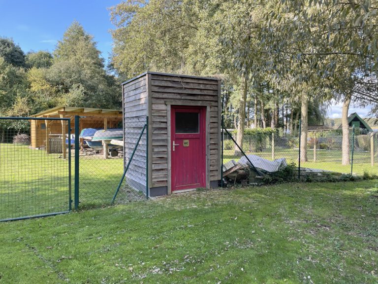 Auf dem Foto ist ein kleines Toilettenhaus LÄRCHE zu erkennen mit eine roten Türe. Das Haus hat links und rechts einen Zaun und dahinter erkennt man Boote unter einem Dach. Der Gras ist grün und deckt den ganzen Boden ab.