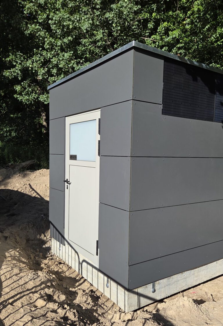 Auf dem Foto ist ein Toilettenhaus PANEEL zu erkennen. Der Boden ist noch eine Baustelle und im Hintergrund sind grüne Baume.