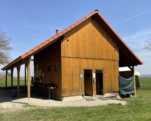 Ein Holzgebäude mit rotem Dach beherbergt die Toiletten für Männer und Frauen. Es ist von Gras umgeben, mit einem Baum und einer offenen Landschaft im Hintergrund.