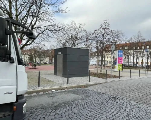 Eine graue Toilettenanlage steht neben einer Kopfsteinpflasterstraße, die von kahlen Bäumen in der ruhigen Stadt gesäumt ist. Links ist ein weißer Lastwagen teilweise sichtbar, während im Hintergrund Reihen von Häusern und bunte Schilder unter einem bewölkten Himmel hervorschauen.