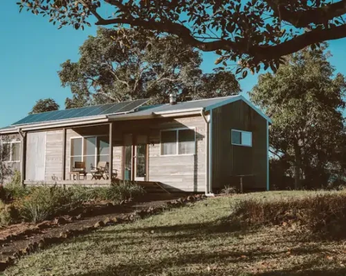 Ein Holzhaus auf einem Grashügel mit Bäumen im Hintergrund unter einem blauen Himmel.