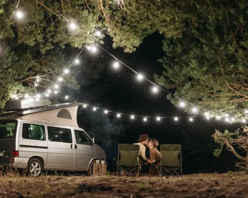 Ein Camper in der Nacht neben einem küssenden Pärchen, umgeben von Bäumen, an denen Lichterketten aufgespannt sind